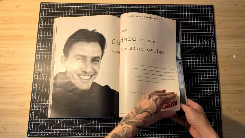 The frame the third eye chose: a magazine open on a black cutting mat, the right page carrying a large typeset German line, an ESPRIT logo at the lower edge, two hands holding the spread flat.