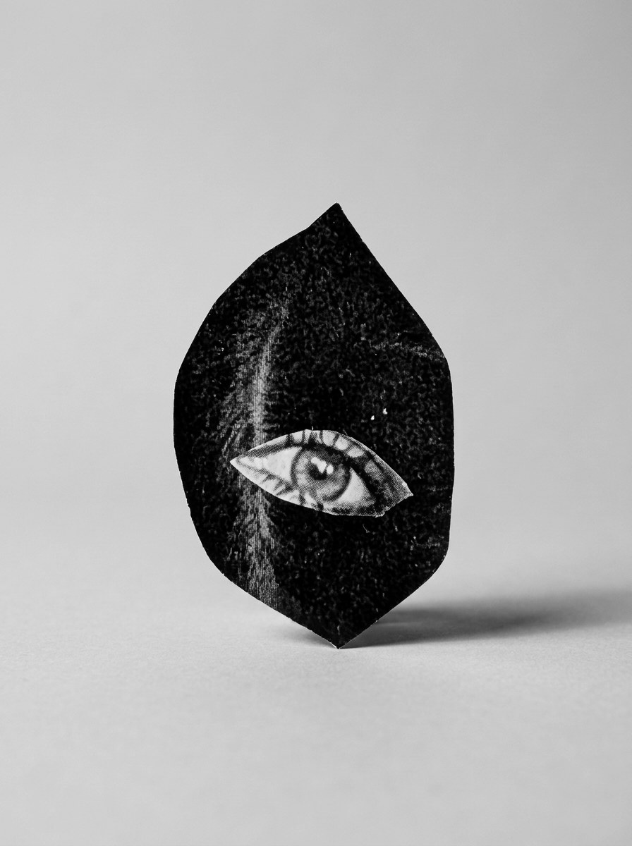 A leaf-shaped piece of cut paper printed with black hair balances on its point against a plain pale grey ground, casting a soft shadow. An eye cut from a magazine is set into it and looks straight out.