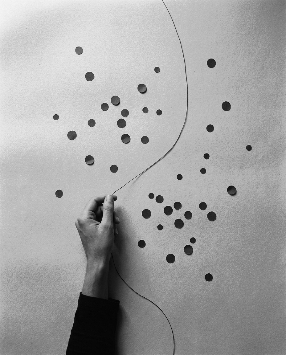 A field of small dark paper circles seen from above, with a hand pulling a single taut thread in a curve across them